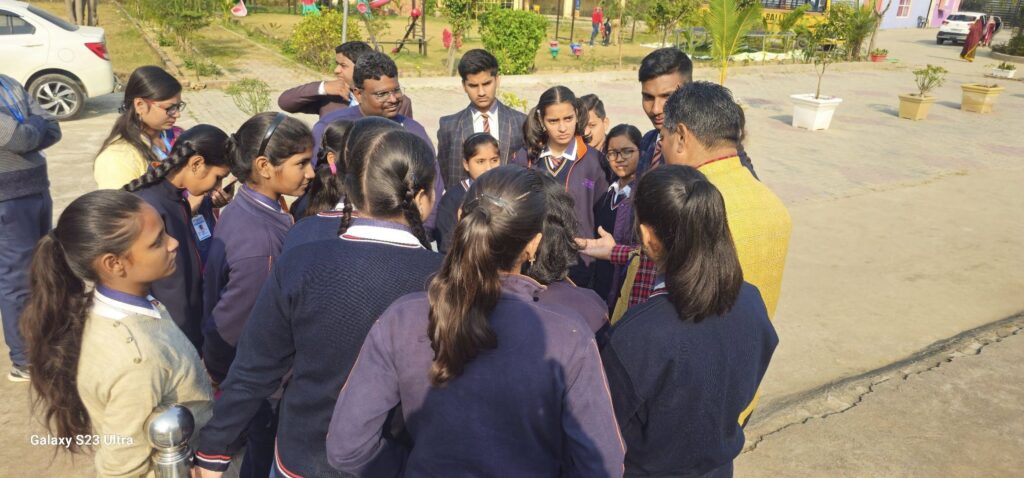 A group of students in navy uniforms stands in a circle outdoors, listening to an adult in a yellow kurta who appears to be talking to them in a schoolyard.",