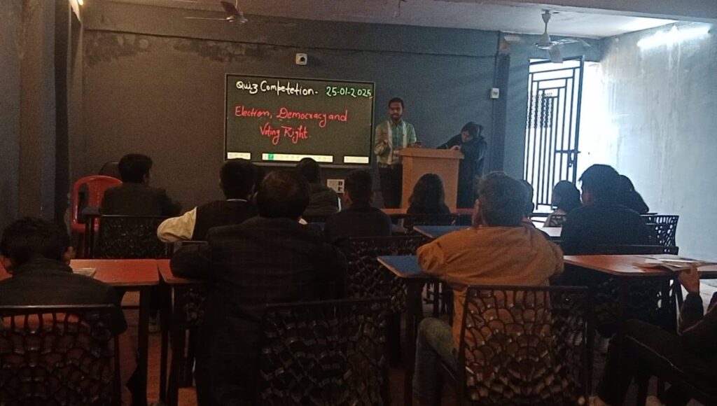Lecture hall scene: a man at a wooden podium presents to students seated at desks; a chalkboard behind shows 'Quiz Competition' and topic 'Election, Democracy and Voting Right'.