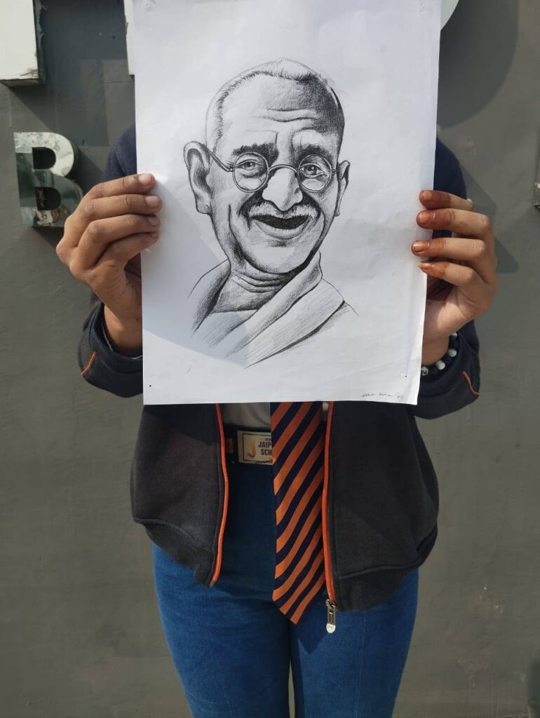 Person wearing a dark jacket and striped tie holds up a large pencil sketch of Mahatma Gandhi in front of their torso against a gray wall.