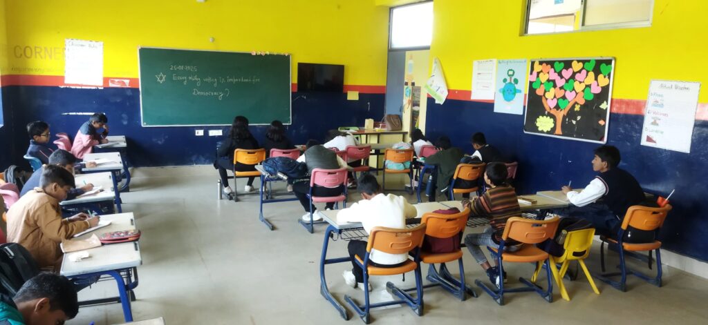 Classroom filled with students seated at desks, writing in notebooks; bright yellow and blue walls, chalkboard at front with notes.