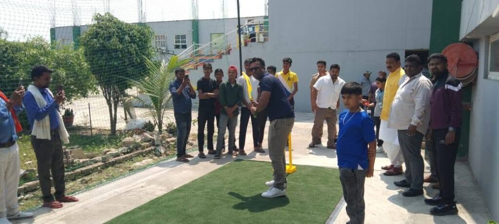 Cricket practice scene: a man with a bat prepares to face a ball on a small turf mat as a group of men and a boy watch outdoors near a sports facility.