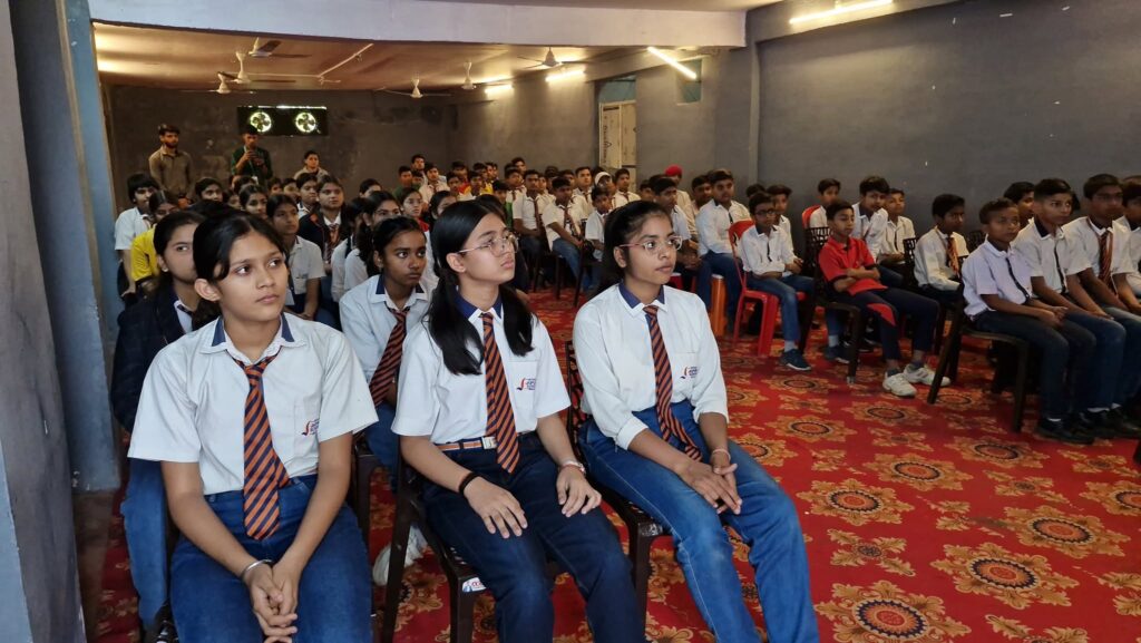 Rows of students in white shirts and orange striped ties sitting in a hall for a school event.